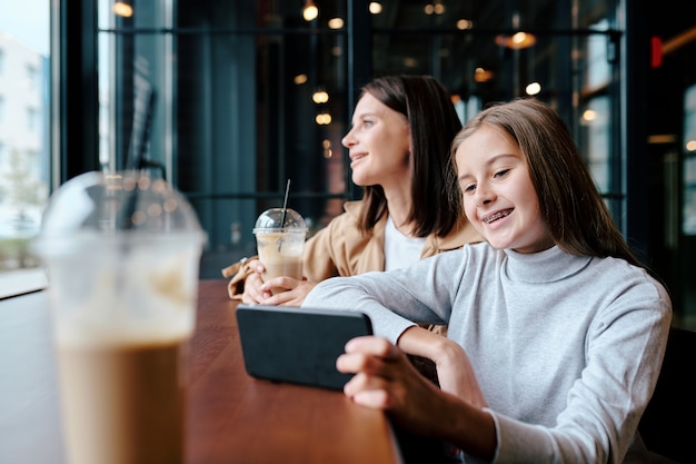 Vrolijk meisje kijken naar grappige video in smartphone zittend in café met haar moeder op achtergrond kijkt door raam