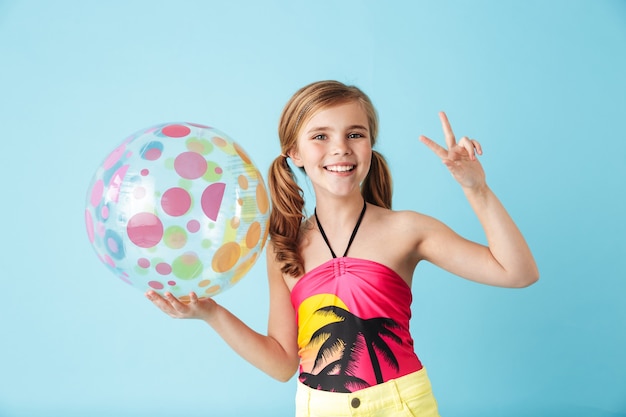 Vrolijk meisje met een zwempak dat geïsoleerd over een blauwe muur staat en een strandbal vasthoudt