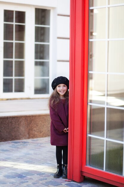 Vrolijk meisje permanent in de buurt van de rode telefooncel in een Bourgondische jas en baret. Londen rode telefooncel. Voorjaar.