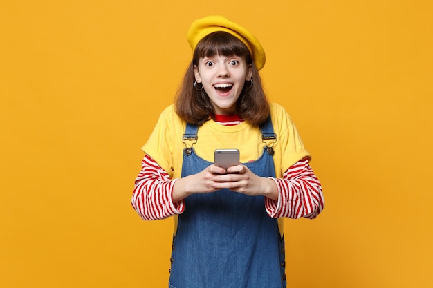 Vrolijk meisje tiener in Franse baret, denim zomerjurk met behulp van mobiele telefoon, sms-bericht typen geïsoleerd op gele achtergrond in studio. Mensen oprechte emoties, lifestyle concept. Bespotten kopie ruimte.