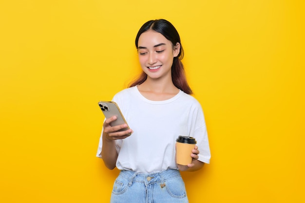 Vrolijk mooi jong meisje in witte t-shirt met koffiekopje en het controleren van nieuwe berichten in smartphone geïsoleerd op gele achtergrond