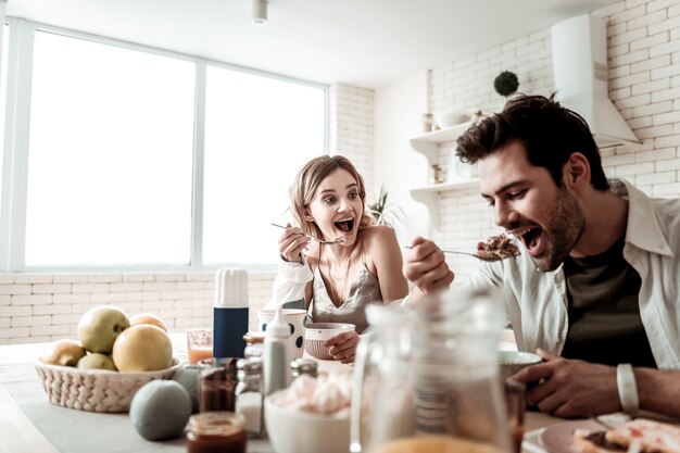 Vrolijk ontbijt. bebaarde knappe positieve man in een wit overhemd voelt zich geweldig tijdens het eten tijdens het ontbijt met zijn vrouw