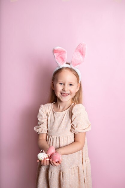 Vrolijk Pasen Een schattig meisje met konijnenoren houdt gekleurde eieren in haar handen en lacht vrolijk roze achtergrond Ruimte voor tekst Foto van hoge kwaliteit