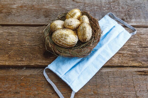 Vrolijk Pasen, voorbereiding op vakantie. Gouden versierde paaseieren in nest met beschermend gezichtsmasker op houten achtergrond