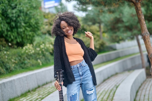 Vrolijk persoon. Vrij slanke Afro-Amerikaanse, vrouw met krullend haar in stijlvolle casual kleding die gitaar aanraakt met de hand die lacht, kijkend naar de camera die overdag in het park staat