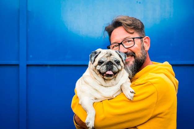 Vrolijk portret van de volwassen man en zijn mooie oude pug dog samen tegen een blauwe muurachtergrond. Concept van geluk en beste vrienden, mensen en dieren levensstijl