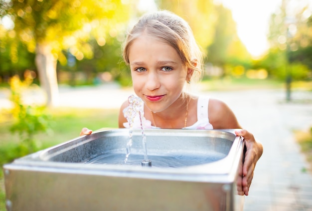 Vrolijk prachtig meisje drinkt koel vers water uit een kleine fontein in een zomers warm zonnig park op een langverwachte vakantie