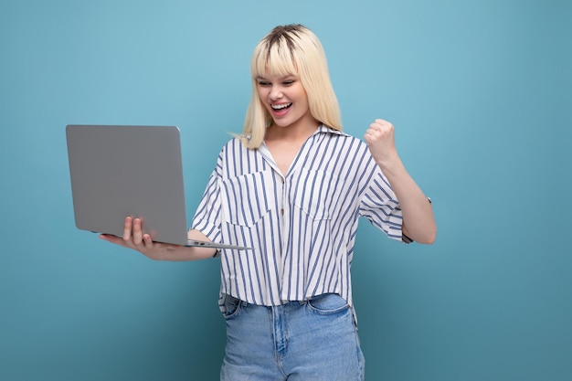 Vrolijk schattig blond jaar oud vrouwelijk persoon in een gestreepte blouse en spijkerbroek met een laptop