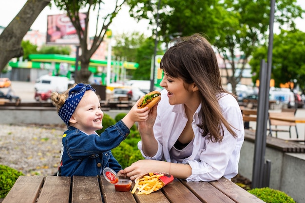 Vrolijk schattig klein meisje zit met haar moeder met hamburger en frietjes op tafel in een café