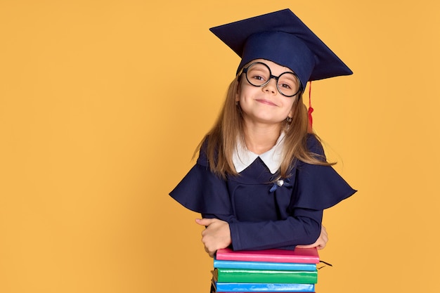 Vrolijk schoolmeisje die in graduatieuitrusting glimlachen terwijl het leunen op stapel van handboeken