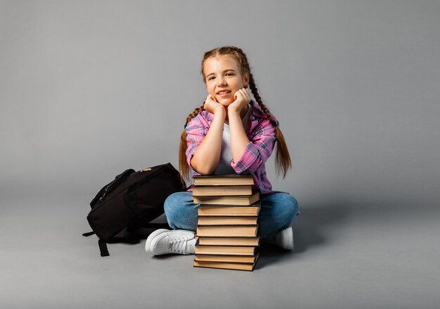 Vrolijk schoolmeisje droomt ervan om in een stapel boeken op een grijze achtergrond in de studio te zitten. Ruimte kopiëren
