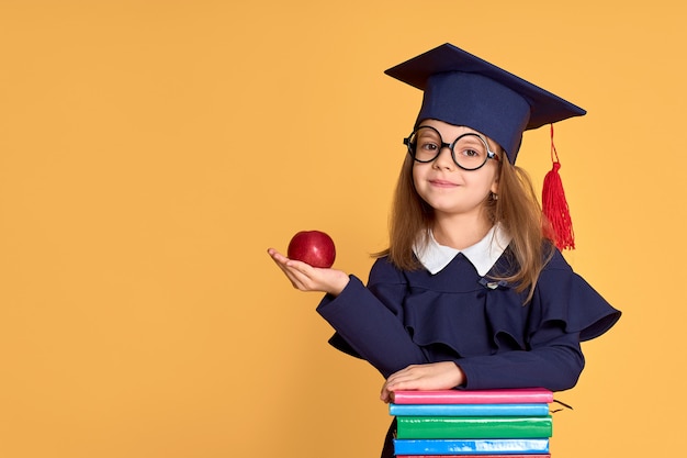 Vrolijk schoolmeisje in de dragende appel van de graduatieuitrusting terwijl status naast stapel van handboeken