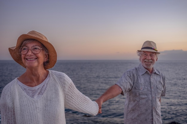 Vrolijk senior koppel met hoeden die genieten van zeevakanties, blanke man en vrouw verliefd bij zonsonderganglicht