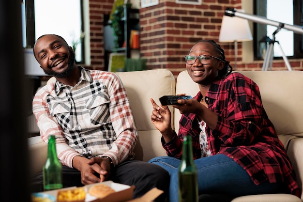 Vrolijk stel dat lacht en samen televisie kijkt, fastfoodmaaltijden eet terwijl vriendin van kanaal wisselt met tv-afstandsbediening. Afhaalmaaltijden en bier om plezier te hebben met film.