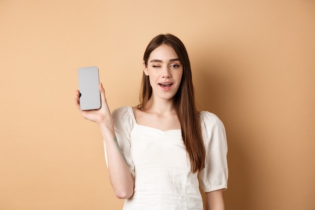 Vrolijk stijlvol meisje toont smartphone-app, toont een leeg telefoonscherm en knipoogt, staande op een beige achtergrond.
