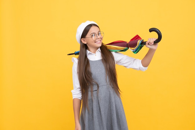 Vrolijk tienerkind houdt kleurrijke parasol vast. kind in baret met regenboogparaplu. herfstseizoen. regenachtig weerbericht. terug naar school. tussen met levendige regenbescherming. gelukkig schoolmeisje in glazen.