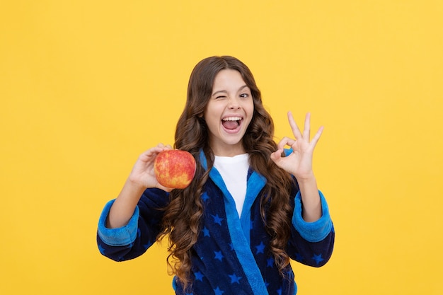 Vrolijk tienermeisje met krullend haar in comfortabele pyjama die een gezonde appel gaat eten met een goed gebaar, op dieet