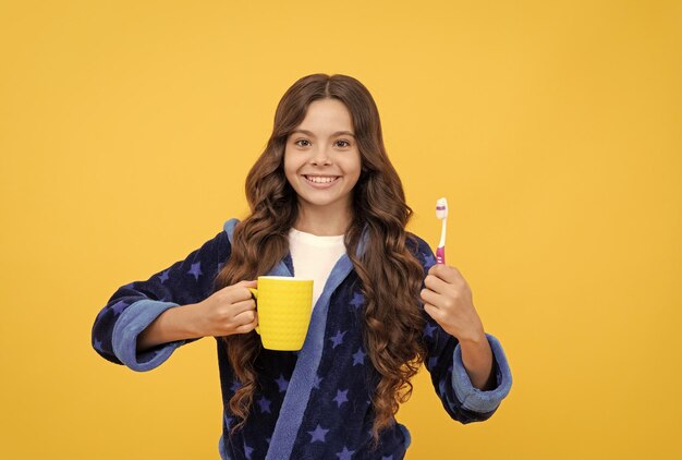 Vrolijk tienermeisje met krullend haar in comfortabele pyjama, tandenborstel en beker hygiënische procedure