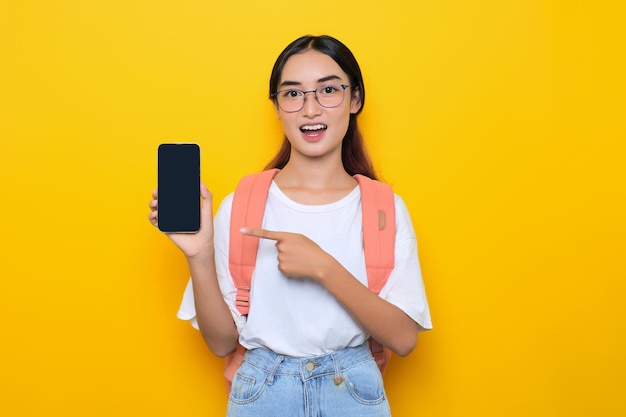 Vrolijk vrij jong studentenmeisje die rugzak en oogglazen dragen die een leeg schermsmartphone tonen die op gele achtergrond wordt geïsoleerd