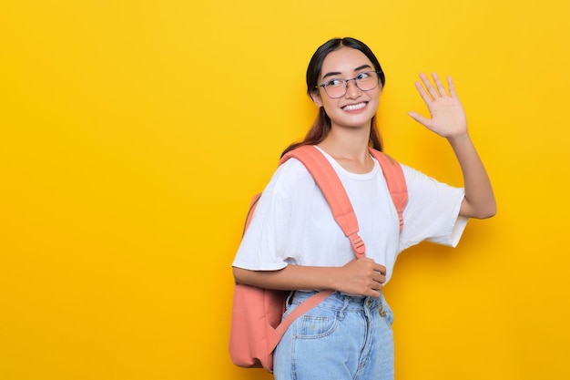 Vrolijk vrij jong studentenmeisje met rugzak en bril die met de hand zwaait, zeg hallo geïsoleerd op gele achtergrond