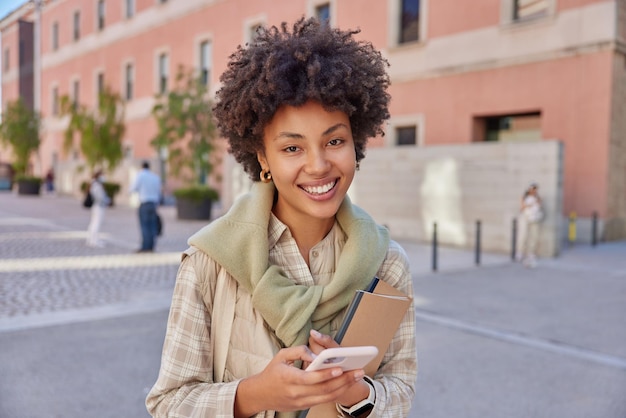 Vrolijk vrouwelijk model met krullend haar controleert e-mail op smartphone draagt notitieblokken gekleed in vrijetijdskleding drukt positieve emoties uit wandelingen buitenshuis tegen stedelijke achtergrond maakt gebruik van mobiele app