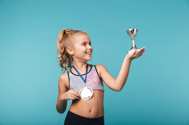 Vrolijk weinig sportenmeisje die de overwinning vieren die over blauwe muur wordt geïsoleerd, die een gouden medaille draagt, een trofee toont