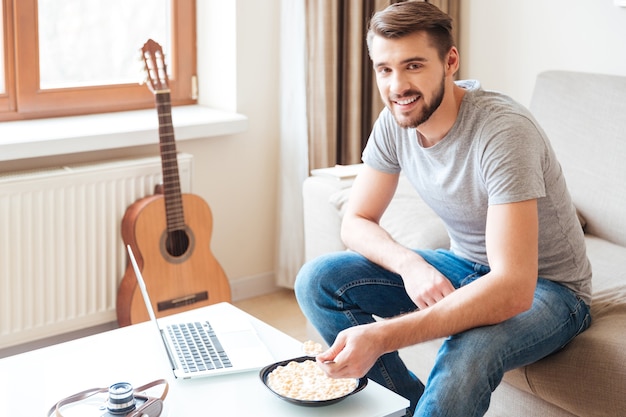 Vrolijke aantrekkelijke bebaarde man met laptop zittend op de bank en ontbijtgranen eten met melk