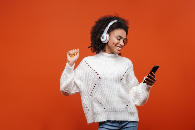 Vrolijke, aantrekkelijke jonge afrikaanse vrouw met een draadloze koptelefoon die geïsoleerd over de rode muur staat, mobiele telefoon vasthoudt, danst