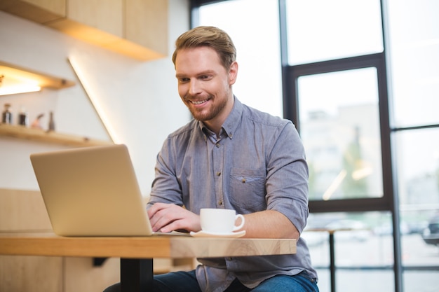 Vrolijke aardige positief opgetogen man aan tafel zitten en op de laptop werken terwijl hij freelance is