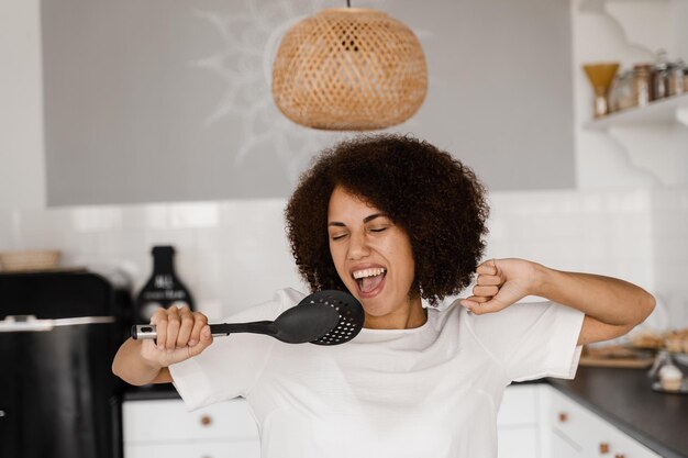 Vrolijke Afrikaanse jonge vrouw die plezier heeft met keukengerei Afro-Amerikaanse huisvrouw in schort zingend met spatels voor het koken microfoon op de keuken