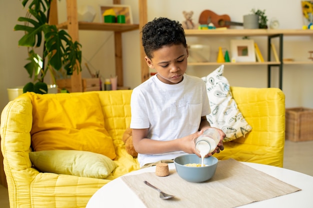 Vrolijke Afrikaanse kleine jongen in t-shirt stukje voedsel in zijn mond zetten zittend op de bank bij tafel in de woonkamer