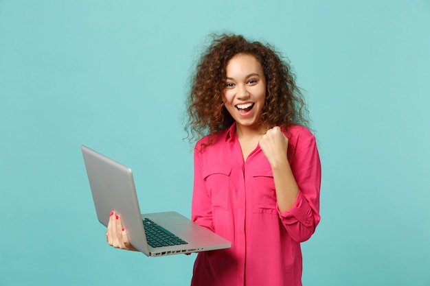 Vrolijke afrikaanse meisje in roze casual kleding winnaar gebaar met behulp van laptop pc-computer geïsoleerd op blauwe turkooizen achtergrond in studio. mensen oprechte emoties levensstijl concept. bespotten kopie ruimte.