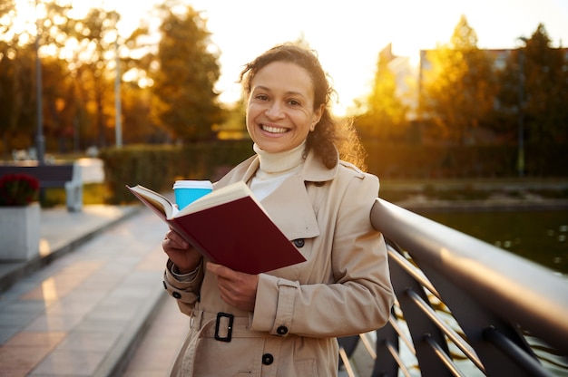 Vrolijke Afrikaanse mooie vrouw geniet van haar weekend buiten, rust in het stadspark op een mooie warme zonnige herfstdag, leest een boek en drinkt koffie, glimlacht terwijl ze naar de camera kijkt