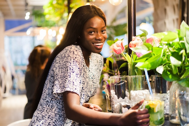Vrolijke Afro-Amerikaanse jonge vrouw in zomerjurk in café is witte bloemen in vaas.