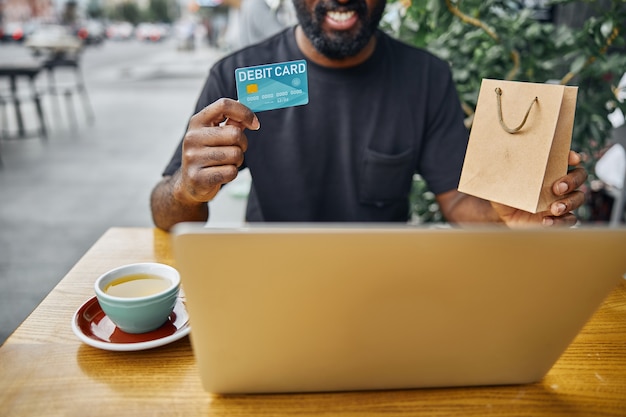 Vrolijke Afro-Amerikaanse man die een papieren zak en een bankpas laat zien terwijl hij voor een laptopscherm zit