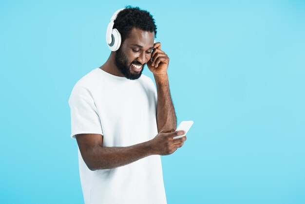 Vrolijke afro-amerikaanse man die muziek luistert met een koptelefoon en een smartphone gebruikt die op blauw is geïsoleerd