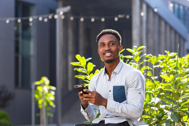 Vrolijke Afro-Amerikaanse man met behulp van mobiele telefoon zittend op een bankje in het stadspark buiten. Gelukkig zwarte man in casual kleding met een smartphone in handen chat op stedelijke moderne achtergrond op straat buiten