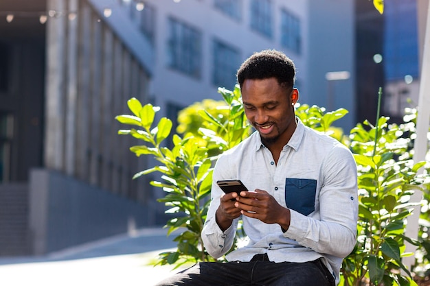 Vrolijke Afro-Amerikaanse man met behulp van mobiele telefoon zittend op een bankje in het stadspark buiten. Gelukkig zwarte man in casual kleding met een smartphone in handen chat op stedelijke moderne achtergrond op straat buiten