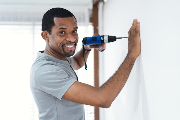 Vrolijke Afro-Amerikaanse manbouwer die een gat boort in een nieuwe witte muur in een nieuw huis met een op batterijen werkende handboor en kijkt naar de camera Volwassene maakt DIY-workshop Verhuizen in nieuw huisconcept
