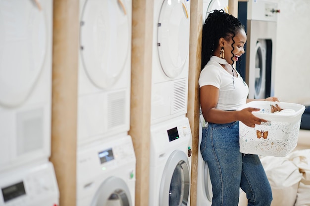 Vrolijke afro-amerikaanse vrouw met witte mand in de buurt van wasmachine in de zelfbedieningswasserij