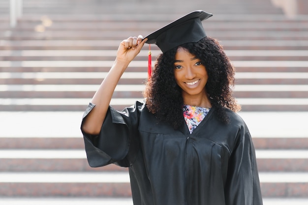Vrolijke Afro-Amerikaanse vrouwelijke afgestudeerde staat voor universiteitsgebouw