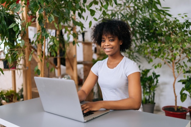 Vrolijke Afro-Amerikaanse vrouwelijke freelancer die op een laptop werkt die aan een bureau zit vanuit een afgelegen kantoor aan huis met een modern biofilie-ontwerp