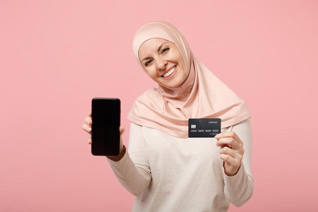 Vrolijke arabische moslimvrouw in hijab lichte kleding poseren geïsoleerd op roze achtergrond. mensen religieuze levensstijl concept. bespotten kopie ruimte. houd mobiele telefoon vast met leeg leeg scherm, creditcard.