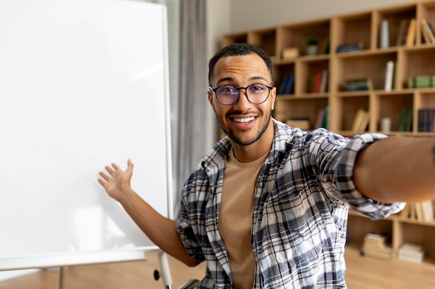 Vrolijke arabische tutor man glimlachend naar de camera en wijzend op whiteboard met online les in de klas