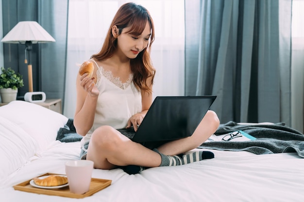 vrolijke aziatische dame die in kleermakerszit op bed zit, eet brood als ontbijt terwijl ze op haar gemak geniet van sociale media-feeds op de computer in een lichte slaapkamer.