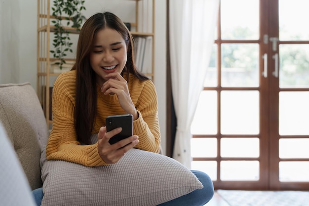 Vrolijke aziatische jonge vrouw die mobiele telefoon gebruikt terwijl ze thuis op een bank zit