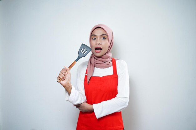 Vrolijke Aziatische moslimvrouw die in rode schort een spatel houdt Kookwaren Kookconcept
