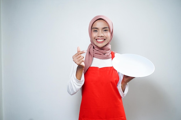 Vrolijke Aziatische moslimvrouw in rode schort met leeg bord Voedsel Advertentie Kokend concept