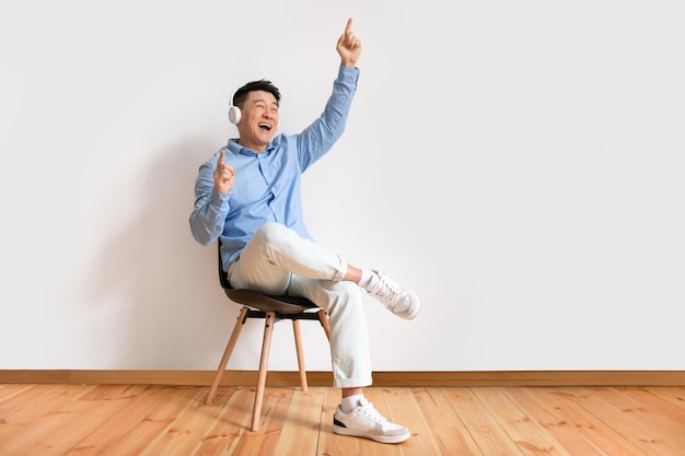 Vrolijke aziatische volwassen man in draadloze koptelefoon zittend op een stoel en luisteren naar muziek tegen witte muur