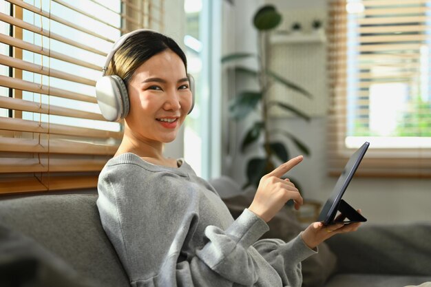 Vrolijke aziatische vrouw die een draadloze hoofdtelefoon draagt en thuis geniet van internetten op een digitale tablet